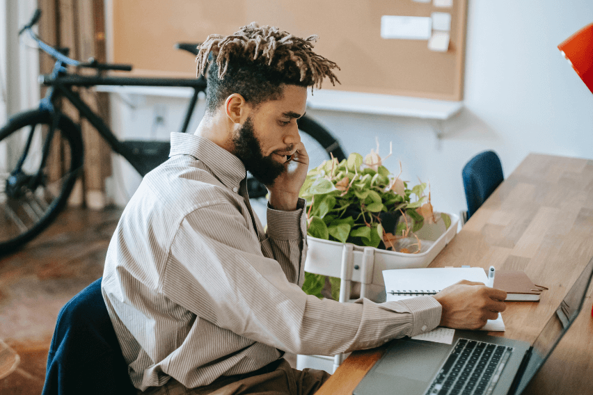 A man sits at a desk with a notepad and laptop while talking on the phone.